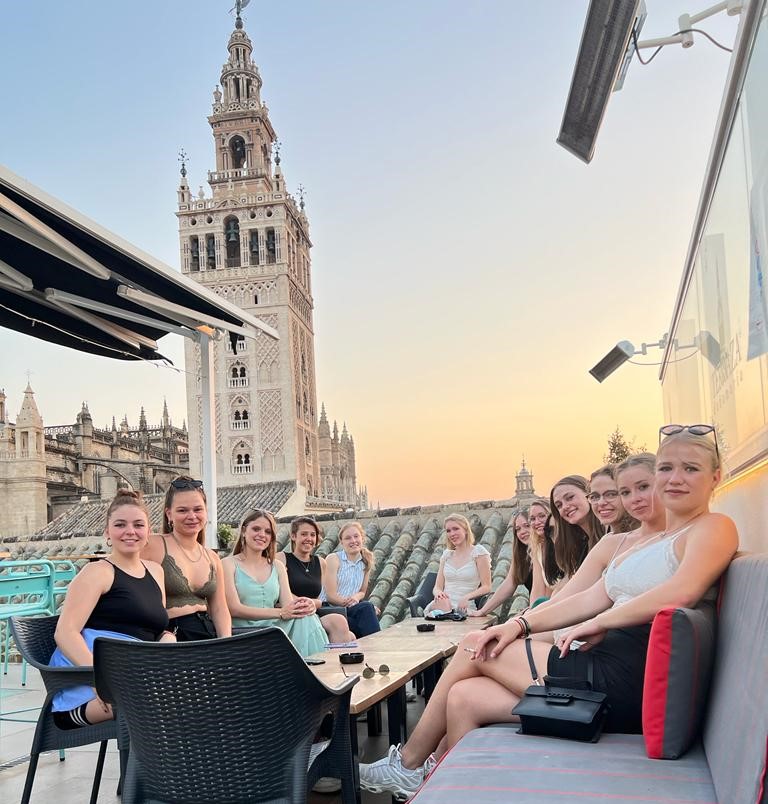 12 junge Frauen sitzen vor einem historischen Turm in der untergehenden Sonne zusammen an einem Tisch.